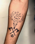 Tattoo of a Viking deer skull with axes and Norse symbols on forearm, showcasing bold ink design.